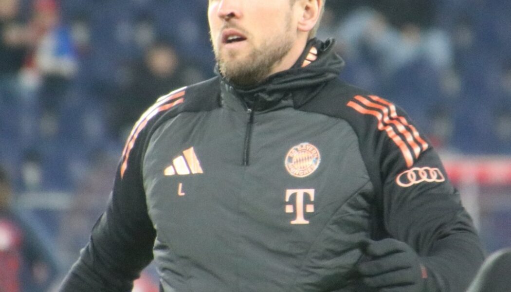 Harry Kane wearing a black Bayern Munich training jacket before a match, with the club badge visible on his chest.